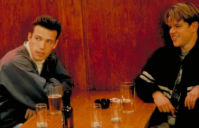 Two men are sitting at a wooden table with drinks and talking in a casual setting.