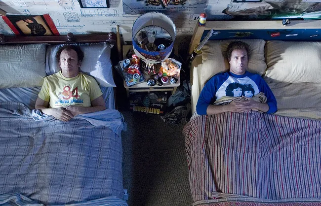 Two men lying in separate beds, looking up at the ceiling, with a cluttered nightstand between them.