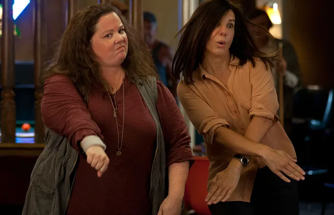 Two women energetically dancing in a bar, one wearing a burgundy top and the other in a beige shirt, with playful expressions.