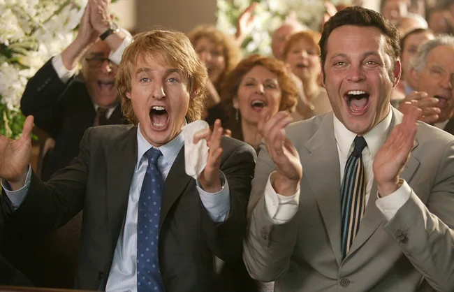 Two men in suits cheering enthusiastically at an event, with a crowd applauding in the background.