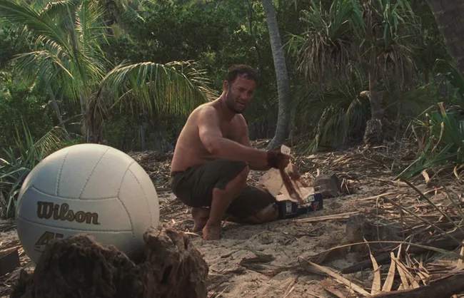Man sits on tropical island beach with a Wilson volleyball, surrounded by palm trees, appearing stranded.
