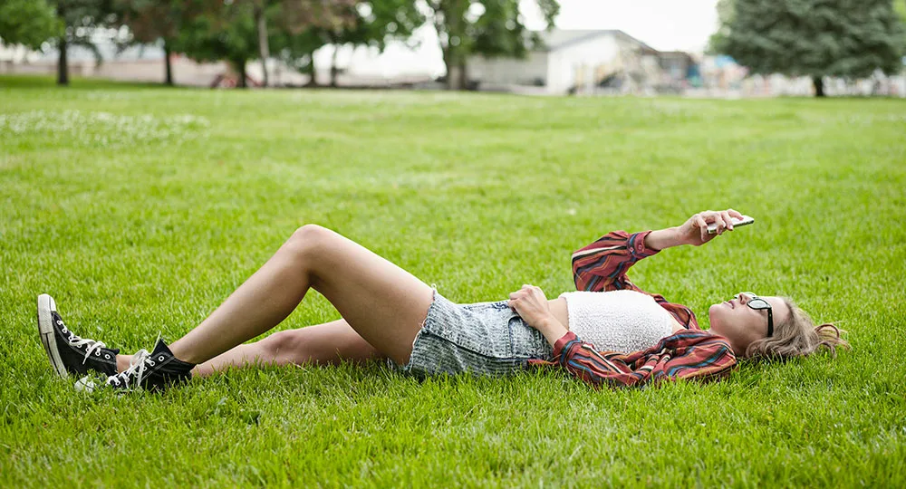 Person lying on grass in a park, looking at a phone, wearing sunglasses, a striped shirt, and denim shorts.