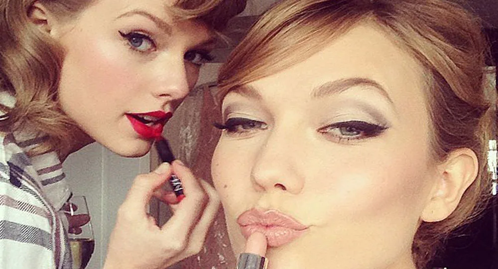 Two women applying lipstick, one in red, the other in nude, posing closely for a selfie.