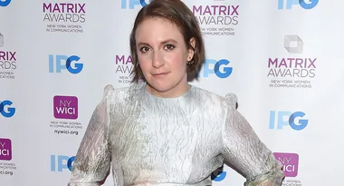A person stands on a red carpet at the Matrix Awards, wearing a silver dress with a backdrop of sponsor logos.