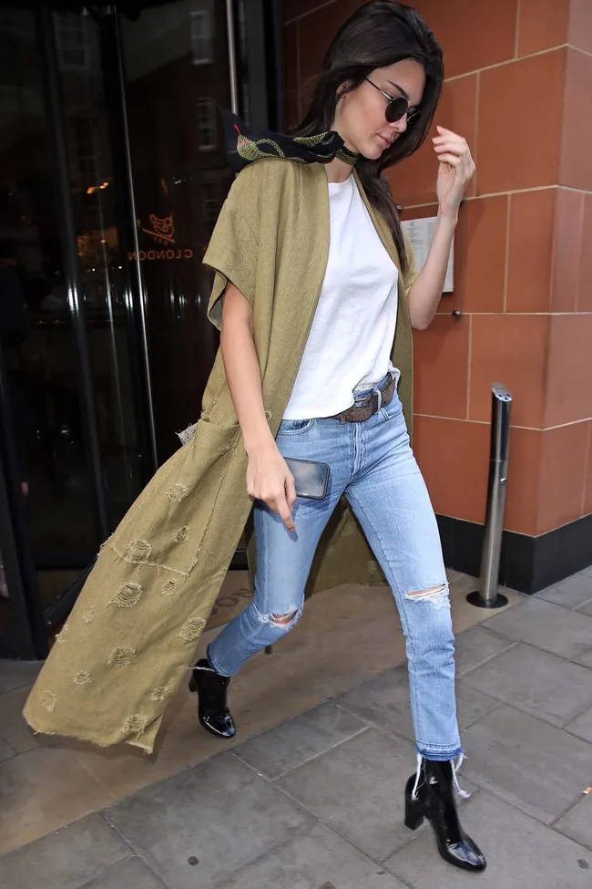 Woman in sunglasses and scarf, wearing a khaki coat, white shirt, distressed jeans, and black boots, walking outside.