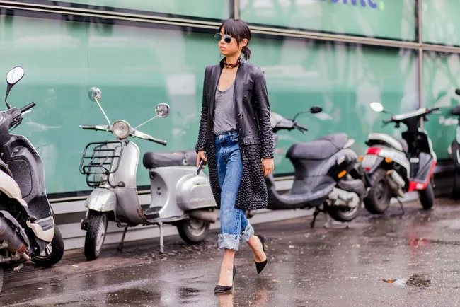 Woman in a studded coat and jeans walks past scooters on a wet street.