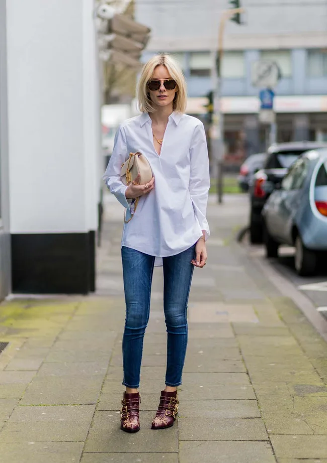 Woman wearing sunglasses, white shirt, skinny jeans, holding a beige clutch, and standing on a city street.