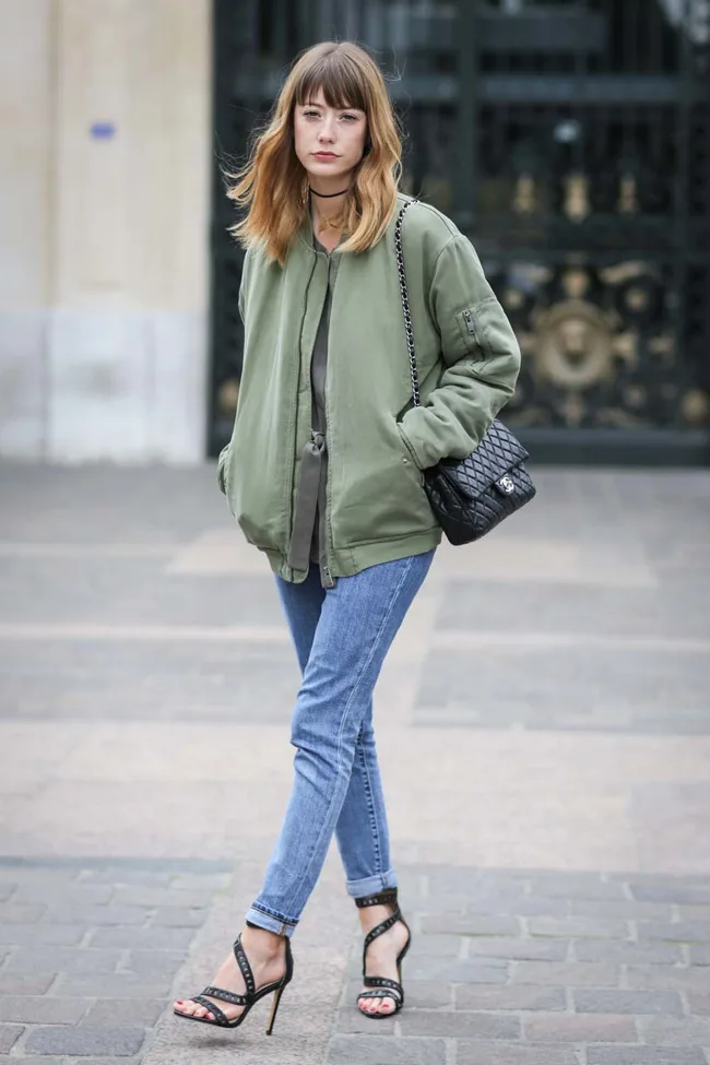 Woman wearing a green bomber jacket, jeans, and heels, carrying a black quilted handbag, standing on a stone path.