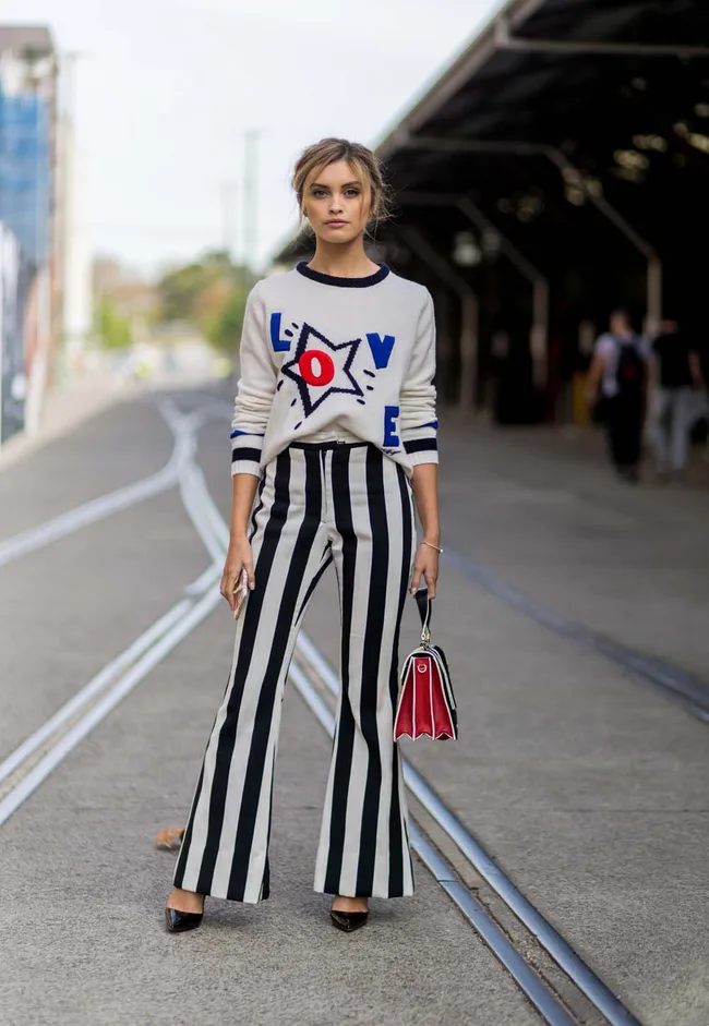 Person wearing a "LOVE" sweater, striped pants, holding a red bag, standing on a street with tram tracks.