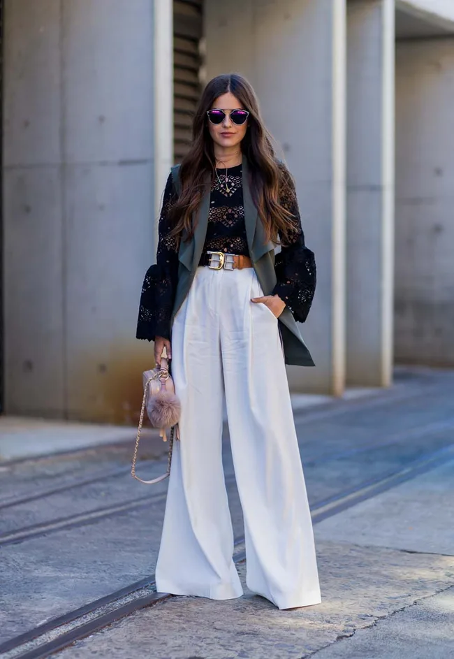 Fashionable woman in a black lace top, white wide-leg pants, sunglasses, holding a pink handbag with a fur pom-pom.