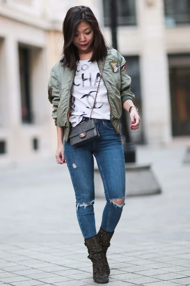 Woman walking in casual outfit, wearing a green jacket, printed tee, ripped jeans, and ankle boots, with a crossbody bag.