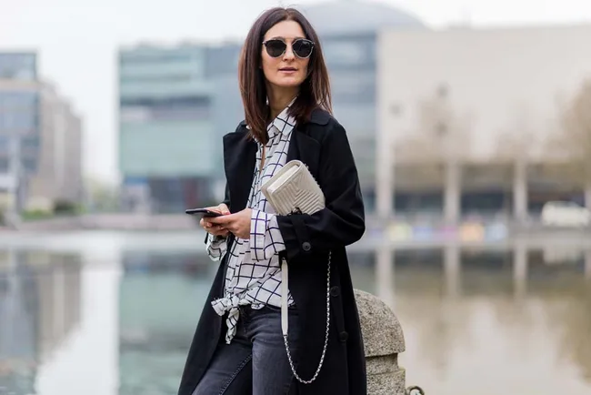 Woman wearing sunglasses and a checkered shirt holds a phone and clutch by a waterfront in an urban area.