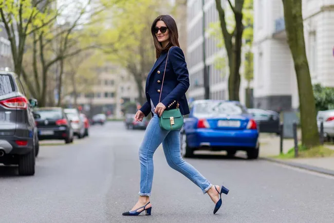 A woman in a blue blazer and jeans walks confidently down a city street, carrying a green handbag.