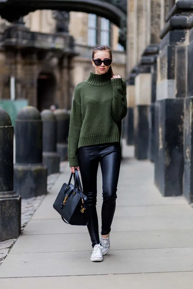 Woman in sunglasses with green sweater, black leather pants, holding black handbag, walking on city street.