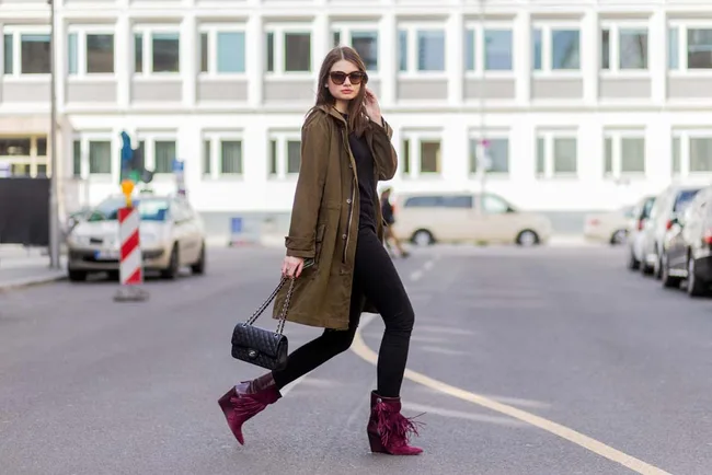 Woman in sunglasses crossing street wearing brown coat, black outfit, and maroon boots, holding a black handbag.