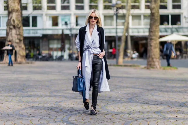 A woman walks confidently on a cobblestone street, wearing sunglasses, a long coat, black pants, and carrying a handbag.