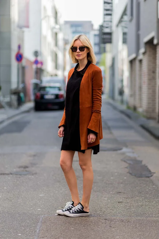 Person in a black dress with brown jacket and sneakers, sunglasses on, standing on a city street.