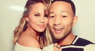 A couple smiling at the camera with light background, the woman has long blonde hair and the man is wearing a striped shirt.