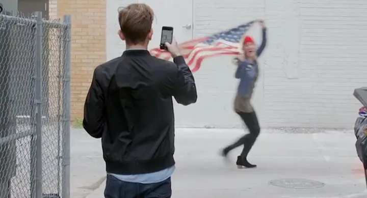 Man photographing woman waving American flag in alley.