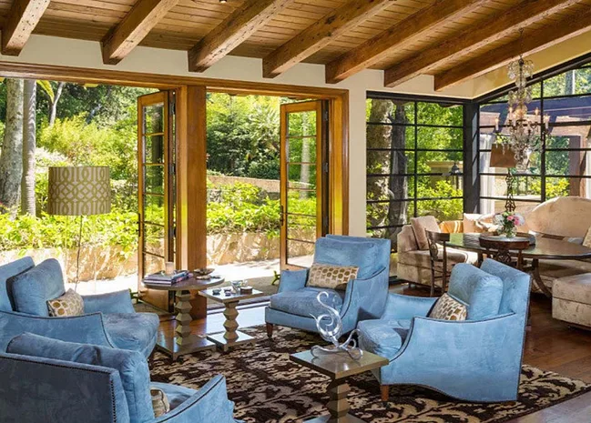 Open living room with blue chairs, wooden beams, and large windows overlooking a garden.