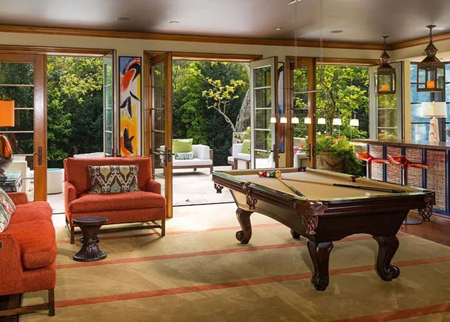 Luxurious living room with a pool table, red chairs, abstract art, and open doors to a lush garden.