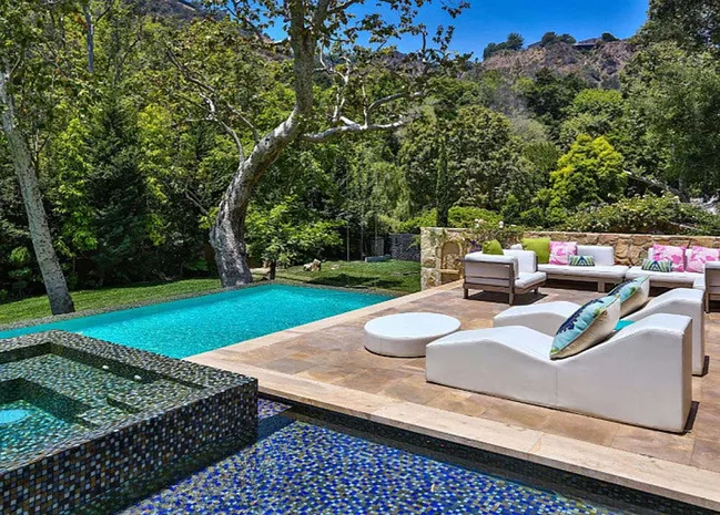 Luxurious backyard with pool, spa, white seating, and vibrant cushions, set against lush greenery and hills.