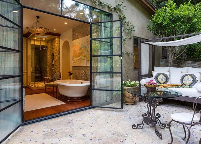 Luxurious bathroom with open glass doors, vintage tub, glass table, and outdoor seating area with lush greenery.