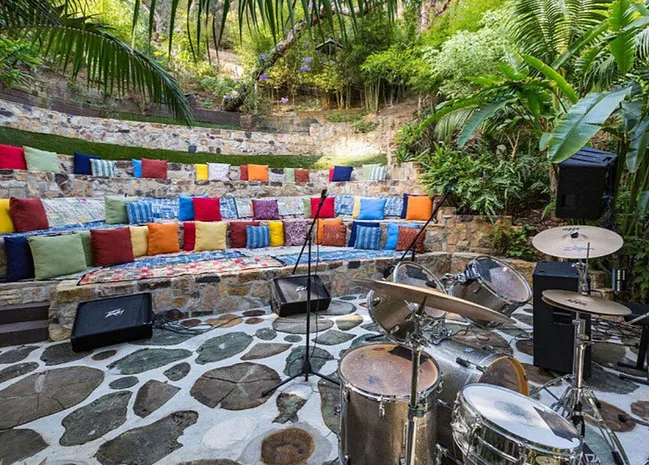 Outdoor amphitheater with colorful cushions, surrounded by greenery, and drum set in foreground.
