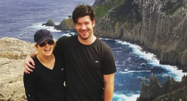 A smiling couple wearing casual clothes poses with a scenic coastal cliff and ocean view in the background.