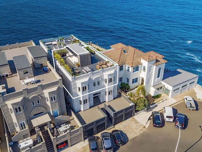 Aerial view of a coastal mansion with multiple levels, rooftop garden, and surrounding ocean. Several cars are parked nearby.