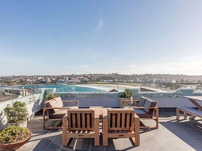 Rooftop patio with wooden furniture overlooking a cityscape and beach under a clear blue sky.