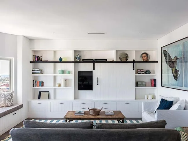 Living room with white built-in shelves, TV, books, pottery, and bird artwork; view of window seat and gray couch.
