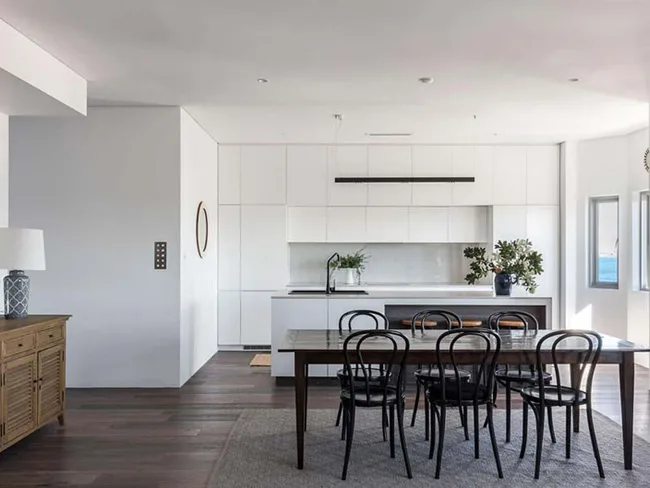 Modern kitchen and dining area with a wooden table, black chairs, white cabinetry, and a sideboard with a lamp.