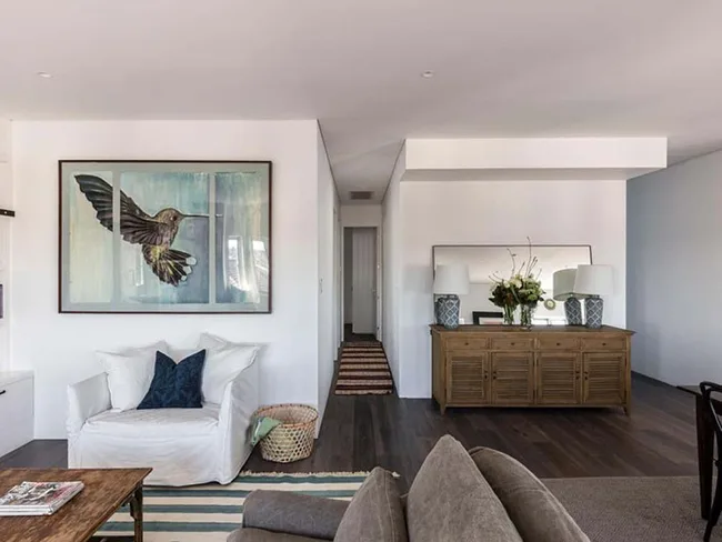 Living room with a white armchair, large hummingbird painting, wooden sideboard, and a mirror with lamps and flowers.