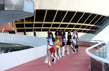Models walking a curved runway outside a modern architectural building, wearing vibrant, geometric-patterned dresses.