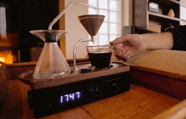 A hand reaches for a cup of coffee from a wooden coffee maker with a digital clock displaying 17:47.