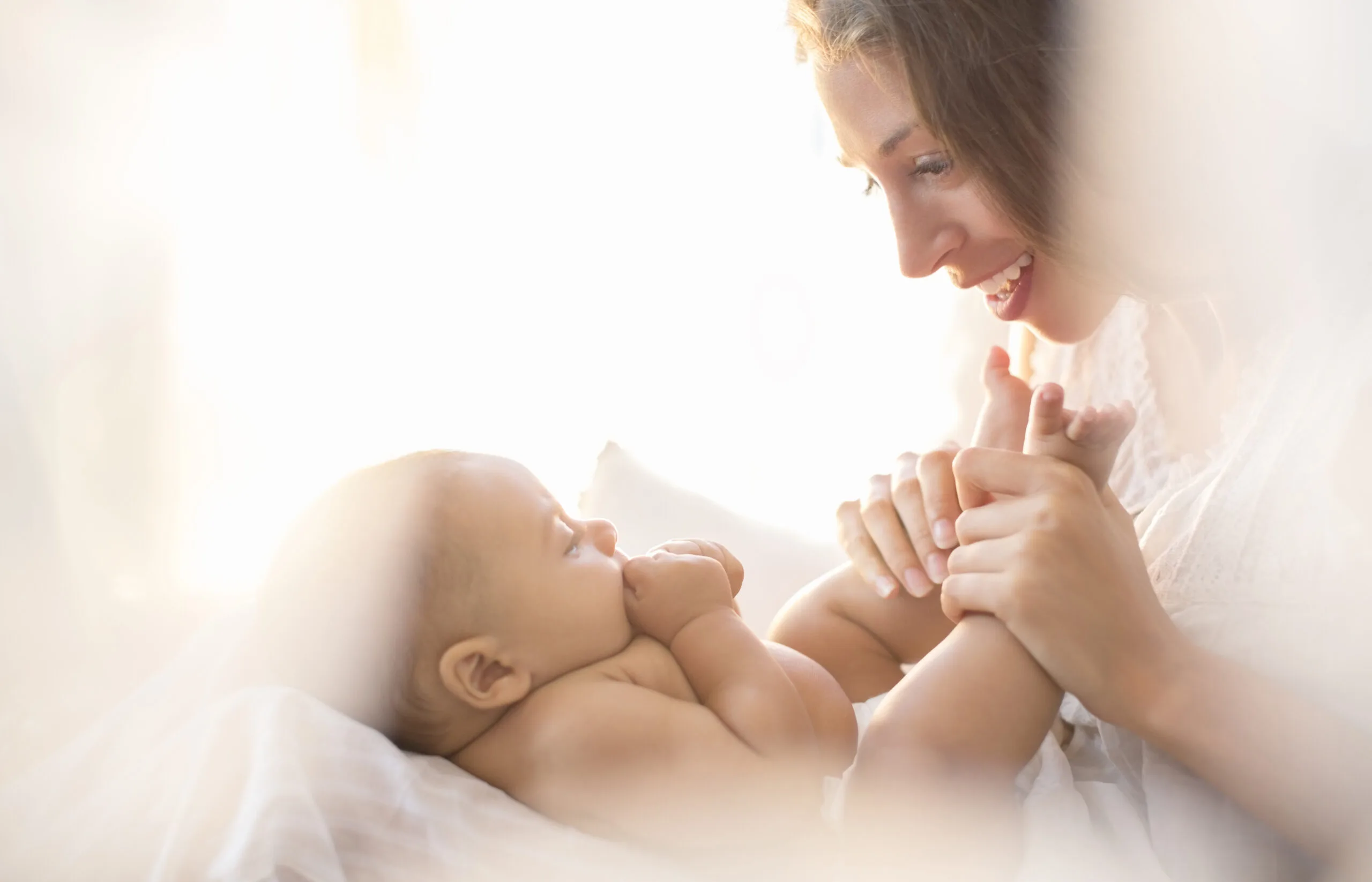 Mother smiling at her baby, holding its feet in a soft, bright setting.