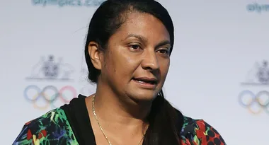 A woman speaking at a podium with Olympic rings in the background.