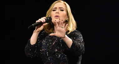 Singer in a sparkly black dress performing on stage, holding a microphone and gesturing with one hand.