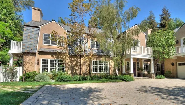 Two-story house with a stone driveway, surrounded by trees and greenery, under a clear blue sky.