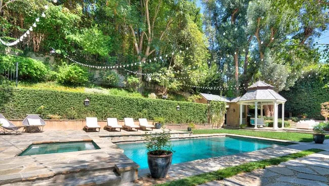 Luxurious backyard with a pool, sun loungers, string lights, and a garden gazebo surrounded by lush greenery.