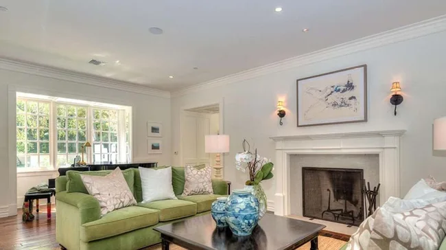 Elegant living room with green sofa, decorative pillows, a fireplace, and artwork above. Large windows with garden view.
