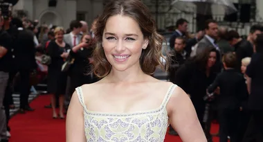 Woman smiling on a red carpet in a white patterned dress, with people in the background.