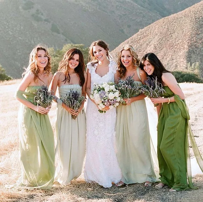 Five women outdoors; four in green bridesmaid dresses, holding flowers, and one in a white wedding gown.