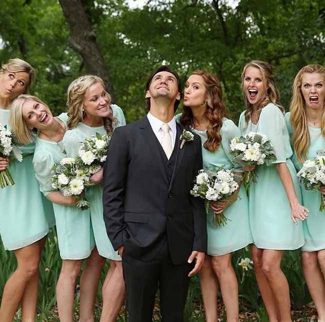 A groom surrounded by bridesmaids in mint dresses making funny faces in a garden setting.