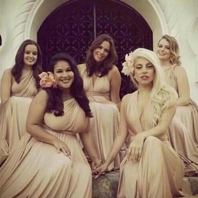 Group of five women in beige dresses, sitting on steps, each with a flower in their hair, smiling for a photo.