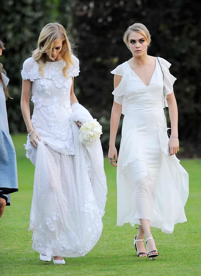 Two women in white dresses walking on grass; one holding a bouquet.