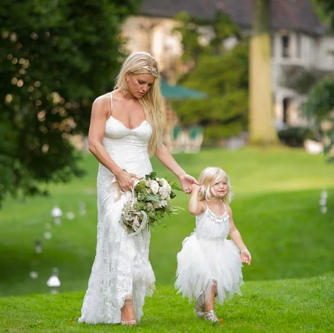 Woman in a white dress holds a bouquet and a toddler's hand as they walk on a grassy field.