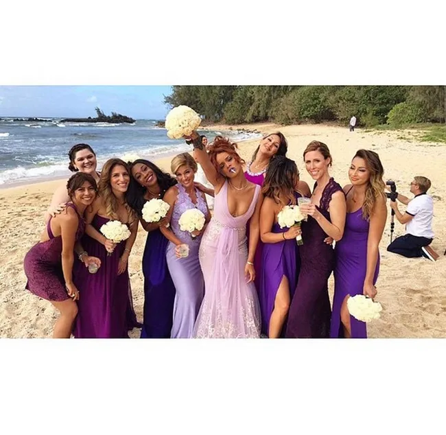 Bridesmaids in purple dresses pose joyfully on a beach with a photographer capturing the moment.