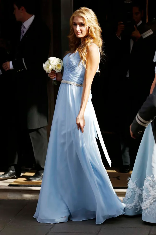 A woman in a light blue gown holds a bouquet of white flowers, standing outside a building.
